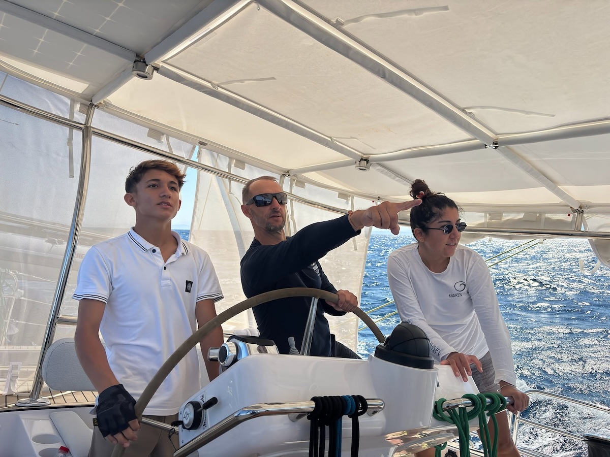 Zwei Crewmitglieder sch&uuml;tzen sich mit lang&auml;rmliger Funktionskleidung. Alle genie&szlig;en das Segeln in der sonnigen &Auml;g&auml;is im Schatten der Pergola